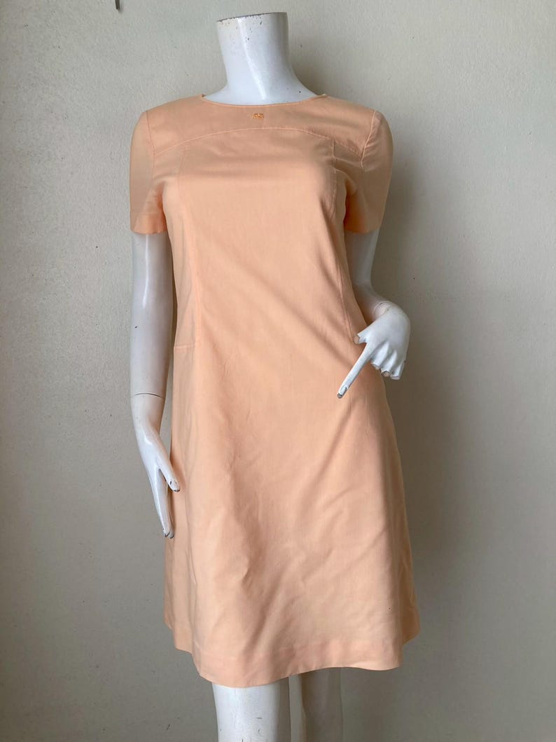 K&ouml;nnte beinhalten: Pfirsichfarbenes Kleid mit kurzen &Auml;rmeln und rundem Ausschnitt. Das Kleid hat ein Knopfdetail am Ausschnitt und eine taillierte Passform. Das Kleid wird auf einer Schaufensterpuppe pr&auml;sentiert.