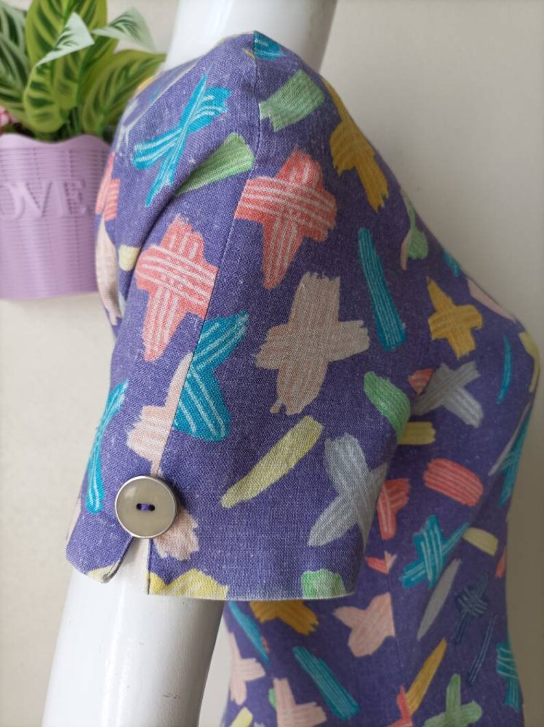 May include: Purple dress with a colorful abstract print. The dress has short sleeves with a button on each cuff.