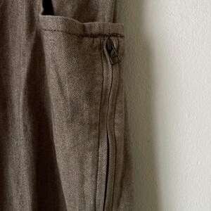 May include: Close-up of a brown herringbone fabric with a zippered pocket. The pocket is on the side of the garment and has a metal zipper. The fabric has a textured appearance.