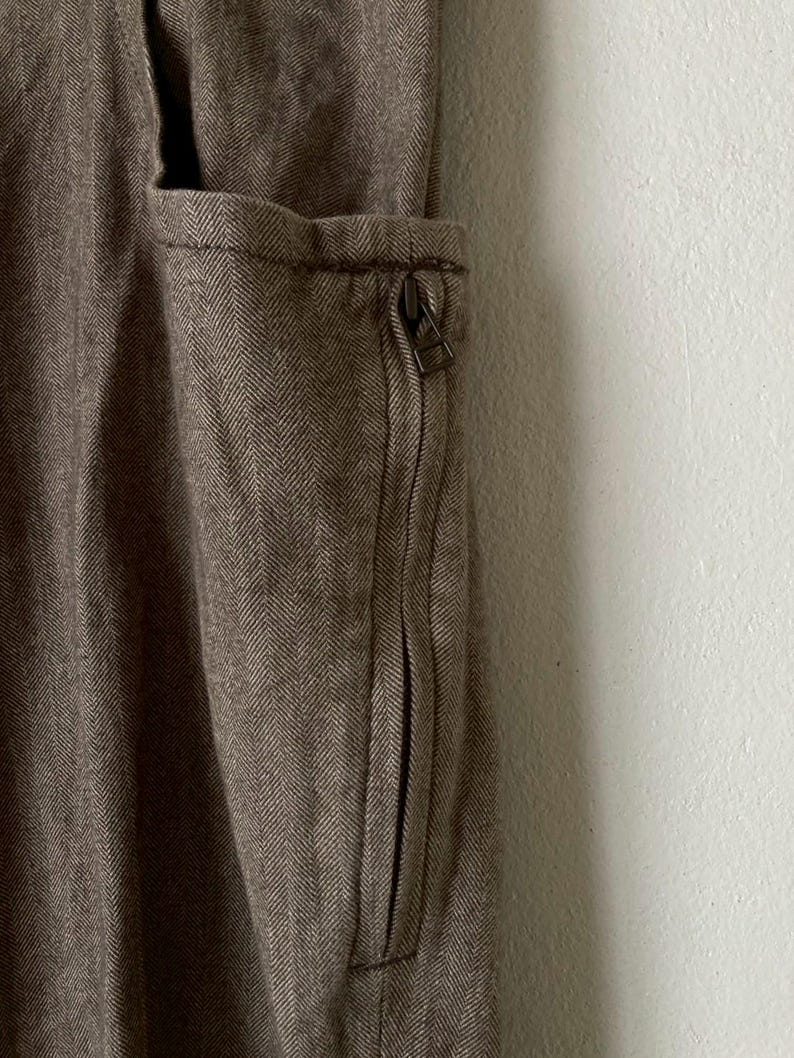 May include: Close-up of a brown herringbone fabric with a zippered pocket. The pocket is on the side of the garment and has a metal zipper. The fabric has a textured appearance.