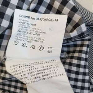 May include: A black and white gingham fabric label with the text "COMME des GAR&Ccedil;ONS Co., Ltd." and care instructions in Japanese and English. The label includes the text "HP-B038", "MADE IN JAPAN", "100% COTTON", "HAND WASH", "DO NOT BLEACH", "DO NOT IRON", "DRY CLEAN", "LINE DRY", "FABRIQUE AU JAPON", "100% COTON", "LAVAGE MAINS", "JAVEL INTERDIT", "NE PAS REPASSER", "NETTOYAGE A SEC", "SECHAGE A L'OMBRE".