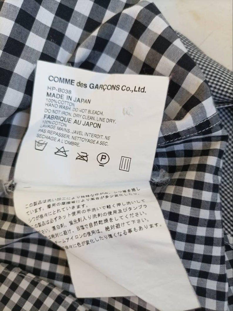 May include: A black and white gingham fabric label with the text "COMME des GAR&Ccedil;ONS Co., Ltd." and care instructions in Japanese and English. The label includes the text "HP-B038", "MADE IN JAPAN", "100% COTTON", "HAND WASH", "DO NOT BLEACH", "DO NOT IRON", "DRY CLEAN", "LINE DRY", "FABRIQUE AU JAPON", "100% COTON", "LAVAGE MAINS", "JAVEL INTERDIT", "NE PAS REPASSER", "NETTOYAGE A SEC", "SECHAGE A L'OMBRE".
