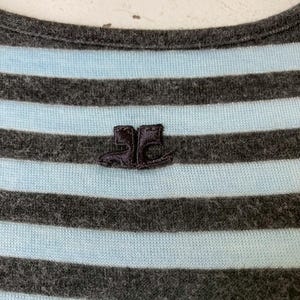Puede incluir: Primer plano de una camiseta a rayas grises y celeste. La camiseta tiene un peque&ntilde;o logotipo bordado de dos botas. El cuello es redondo. La tela parece suave y c&oacute;moda.