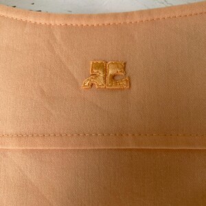 K&ouml;nnte beinhalten: Nahaufnahme eines pfirsichfarbenen Stoffes mit einem kleinen, gestickten Logo in der oberen rechten Ecke. Das Logo ist ein stilisiertes Design in einem passenden Pfirsichton. Der Stoff hat eine subtile Textur und eine sichtbare Naht.