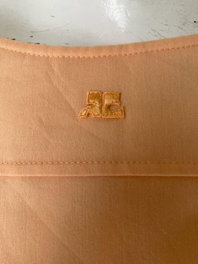 K&ouml;nnte beinhalten: Nahaufnahme eines pfirsichfarbenen Stoffes mit einem kleinen, gestickten Logo in der oberen rechten Ecke. Das Logo ist ein stilisiertes Design in einem passenden Pfirsichton. Der Stoff hat eine subtile Textur und eine sichtbare Naht.