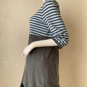 Puede incluir: Una blusa a rayas grises y azul claro con mangas tres cuartos. La blusa tiene una secci&oacute;n inferior gris lisa y se muestra en un maniqu&iacute;. La parte inferior de la blusa se combina con una falda negra.