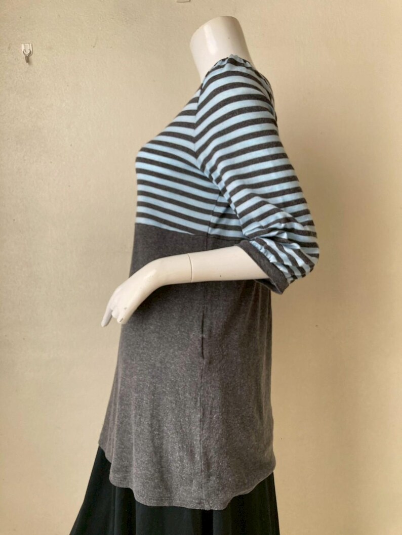 Puede incluir: Una blusa a rayas grises y azul claro con mangas tres cuartos. La blusa tiene una secci&oacute;n inferior gris lisa y se muestra en un maniqu&iacute;. La parte inferior de la blusa se combina con una falda negra.