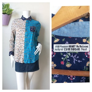 May include: A blue and beige long-sleeved button-up shirt with a floral pattern. The shirt has a patch pocket on the left side and a denim collar. The shirt has a label that reads "1989 Pleasant HEART Be Relieved natural CUBE SUGAR trust".
