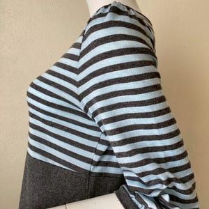 Puede incluir: Una blusa de manga larga a rayas grises y celeste. La blusa presenta un corpi&ntilde;o y mangas grises con rayas horizontales celeste y grises. Las mangas tienen un detalle fruncido en el hombro y un pu&ntilde;o gris.
