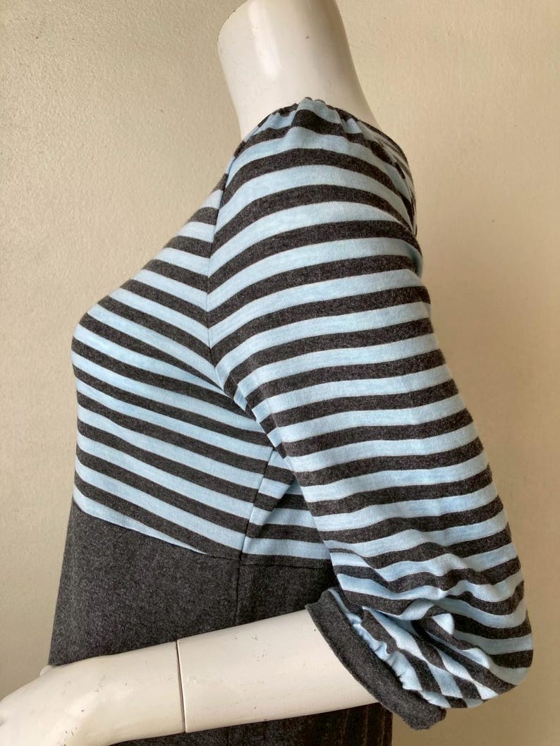 Puede incluir: Una blusa de manga larga a rayas grises y celeste. La blusa presenta un corpi&ntilde;o y mangas grises con rayas horizontales celeste y grises. Las mangas tienen un detalle fruncido en el hombro y un pu&ntilde;o gris.