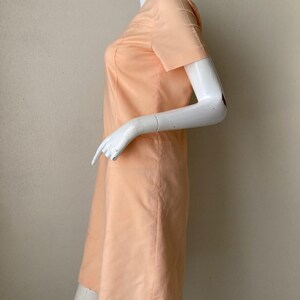 K&ouml;nnte beinhalten: Ein pfirsichfarbenes Kleid mit kurzen &Auml;rmeln. Das Kleid hat eine einfache, gerade Silhouette und einen bescheidenen Ausschnitt. Der Stoff scheint ein glattes, einfarbiges Material zu sein. Das Kleid wird auf einer Schaufensterpuppe pr&auml;sentiert.