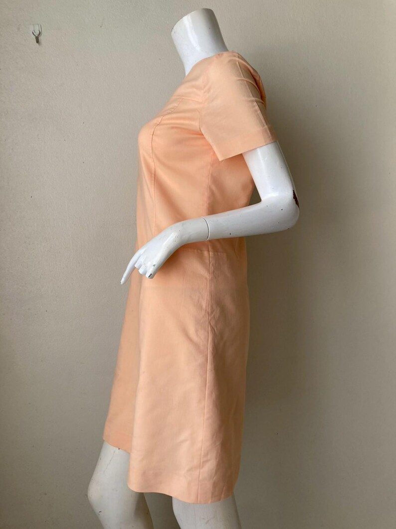 K&ouml;nnte beinhalten: Ein pfirsichfarbenes Kleid mit kurzen &Auml;rmeln. Das Kleid hat eine einfache, gerade Silhouette und einen bescheidenen Ausschnitt. Der Stoff scheint ein glattes, einfarbiges Material zu sein. Das Kleid wird auf einer Schaufensterpuppe pr&auml;sentiert.