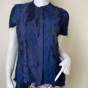 Puede incluir: Una camisa azul de manga corta con un diseño de patchwork. La camisa tiene un frente abotonado y un dobladillo ligeramente asimétrico.