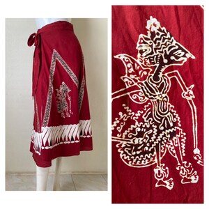 May include: A red wrap skirt with a white batik pattern featuring a traditional Indonesian shadow puppet design. The skirt has a tie closure at the waist.