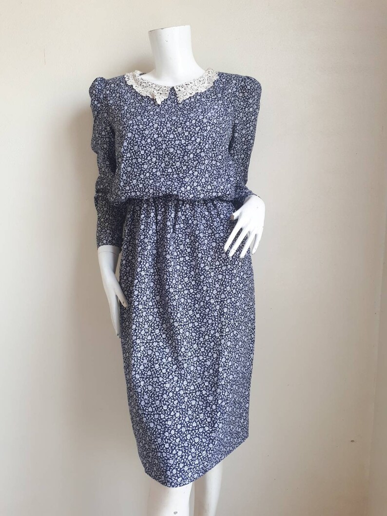 May include: A blue and white floral dress with a white lace collar. The dress has long sleeves and a fitted bodice.