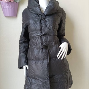 May include: A dark grey, quilted winter coat with a large collar and toggle fastenings. The coat is displayed on a mannequin and has long sleeves. The coat appears to be made of a lightweight material and is knee-length.