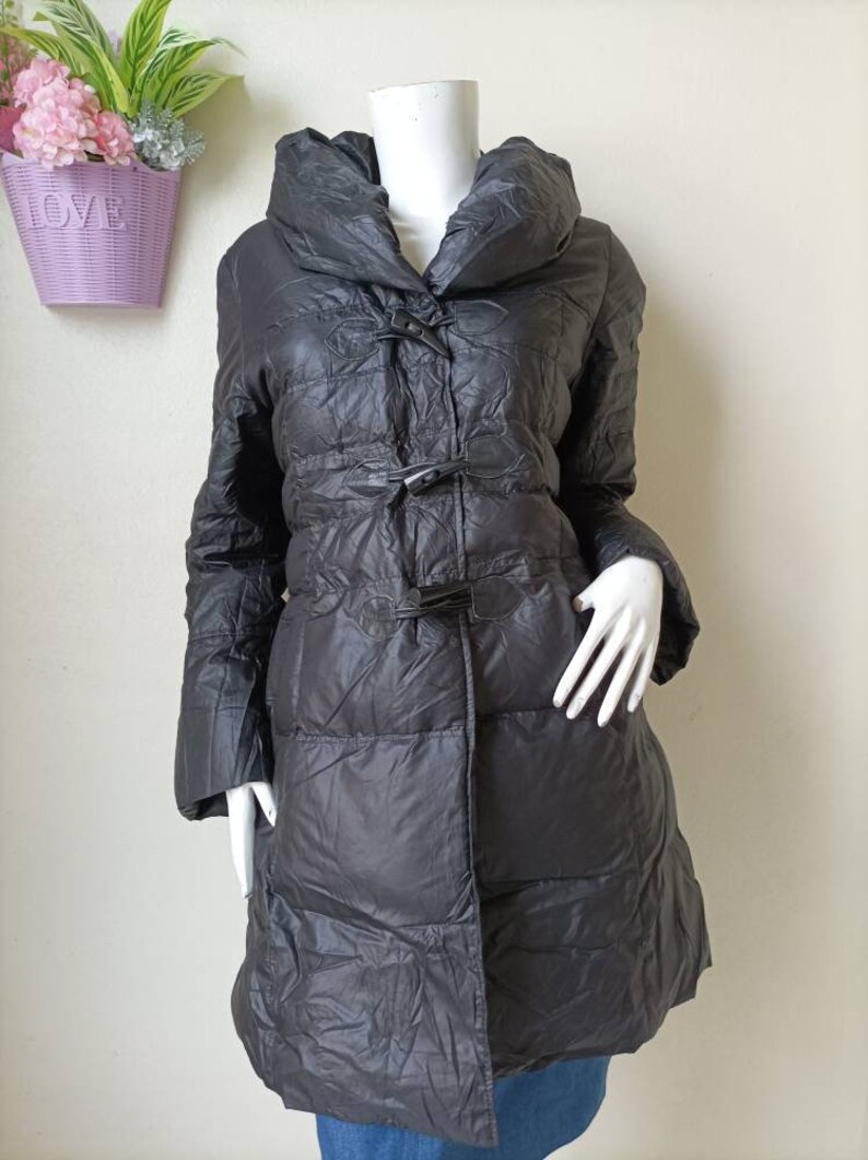 May include: A dark grey, quilted winter coat with a large collar and toggle fastenings. The coat is displayed on a mannequin and has long sleeves. The coat appears to be made of a lightweight material and is knee-length.