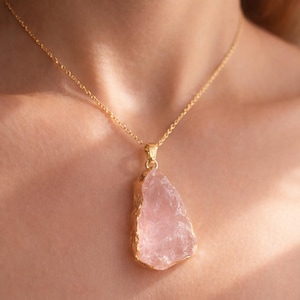 May include: A gold necklace with a pendant. The pendant is a large, irregular-shaped, pale pink gemstone with gold edging. The necklace is displayed on a person's chest.