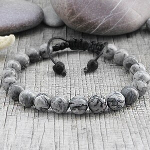 May include: A gray and black stone beaded bracelet with a black cord closure. The bracelet is on a wooden surface.