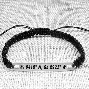 May include: Black braided bracelet with a silver tag engraved with the coordinates "39.0416&deg; N, 94.5922&deg; W".