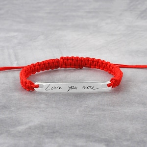May include: Red string bracelet with a silver bar charm engraved with the handwritten message "Love you more".