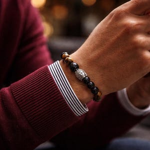 May include: A beaded bracelet featuring tiger's eye and black onyx beads, with a silver Buddha head charm. The bracelet is worn on a wrist, with a burgundy long-sleeve shirt and a white and navy striped cuff.