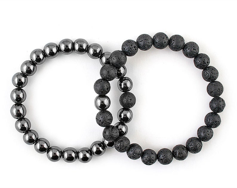 May include: Two beaded bracelets. One bracelet features polished, metallic gray beads. The other bracelet is made of porous, black lava rock beads. Both bracelets are circular and sit on a white background.