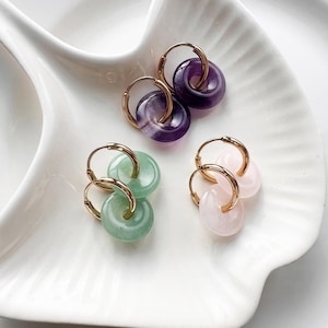 Puede incluir: Tres pares de pendientes de aro con colgantes de piedras preciosas. Los pendientes presentan aros dorados y colgantes en tonos morado, verde y rosa. Los pendientes se exhiben en un plato blanco decorativo.