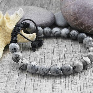 May include: A gray and black beaded bracelet with a black cord closure. The bracelet is made of round, smooth beads with a marbled pattern. The bracelet is lying on a wooden surface with a starfish and stones.