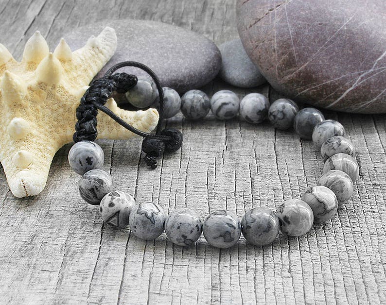 May include: A gray and black beaded bracelet with a black cord closure. The bracelet is made of round, smooth beads with a marbled pattern. The bracelet is lying on a wooden surface with a starfish and stones.