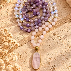 May include: A multi-strand beaded necklace featuring translucent iridescent beads, deep purple amethyst-colored beads, and pale pink rose quartz beads. The necklace includes a large, oval-shaped rose quartz pendant set in a gold-toned metal frame.