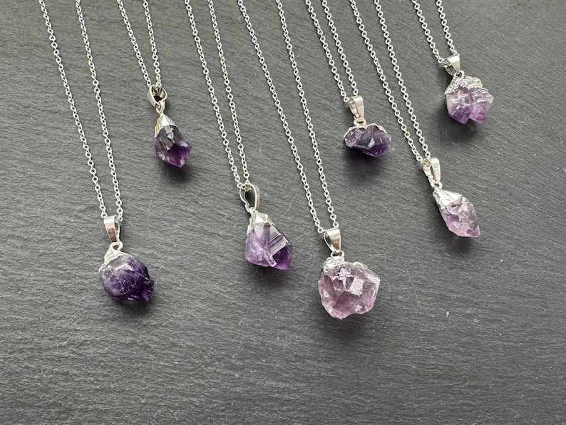 May include: Seven silver necklaces with amethyst crystal pendants. The amethyst crystals are all different shapes and sizes, and they have a purple and light purple color.