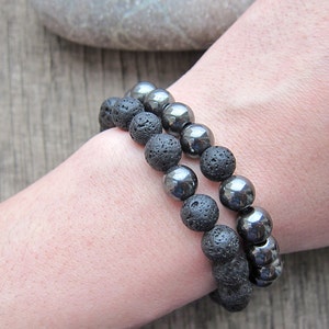 May include: Two beaded bracelets. One bracelet features porous black lava rock beads, and the other has shiny, metallic hematite beads. The bracelets are worn on a wrist, showcasing the contrast in textures and colors.