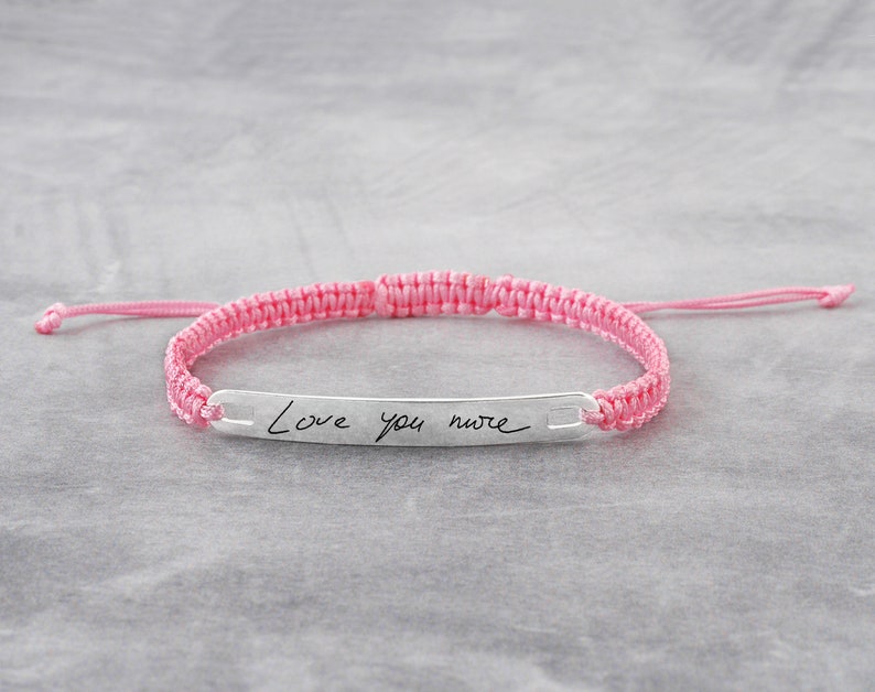 May include: A pink braided bracelet with a silver bar charm engraved with the handwritten message "Love you more".