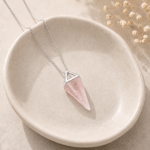 Peut inclure: Un collier pendentif composé d'un cristal de quartz rose facetté en forme de cône pointu. Le cristal est maintenu par une bélière métallique triangulaire argentée et est suspendu à une fine chaîne à maillons de couleur argentée.