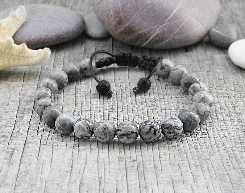 May include: A gray and black stone beaded bracelet with a black cord closure. The bracelet is on a wooden surface.