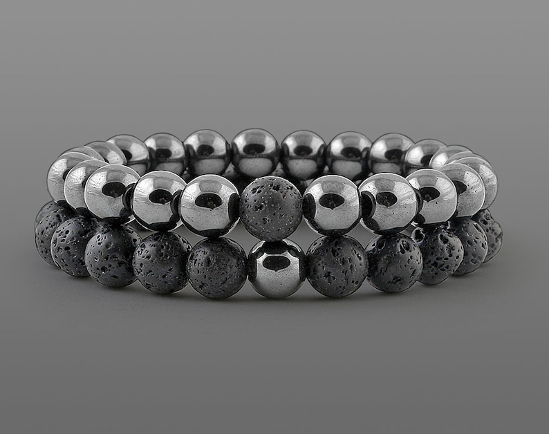 May include: Two beaded bracelets. The top bracelet features polished, silver-colored beads. The bottom bracelet has dark gray, textured lava stone beads. The bracelets are stacked on a gray background.
