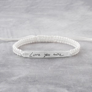 May include: White braided bracelet with a silver bar charm engraved with the handwritten message "Love you more".