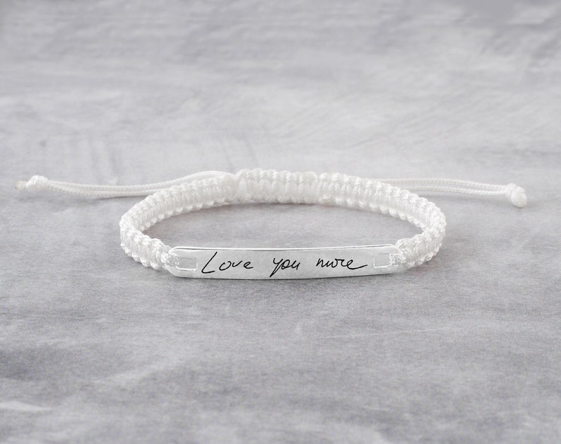May include: White braided bracelet with a silver bar charm engraved with the handwritten message "Love you more".
