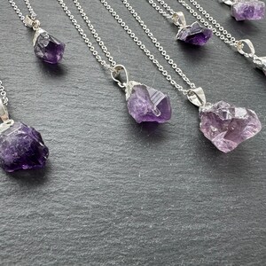 May include: A set of seven silver necklaces with amethyst crystal pendants. The crystals are raw and have a natural, irregular shape. The necklaces are all different lengths and the crystals are various sizes.