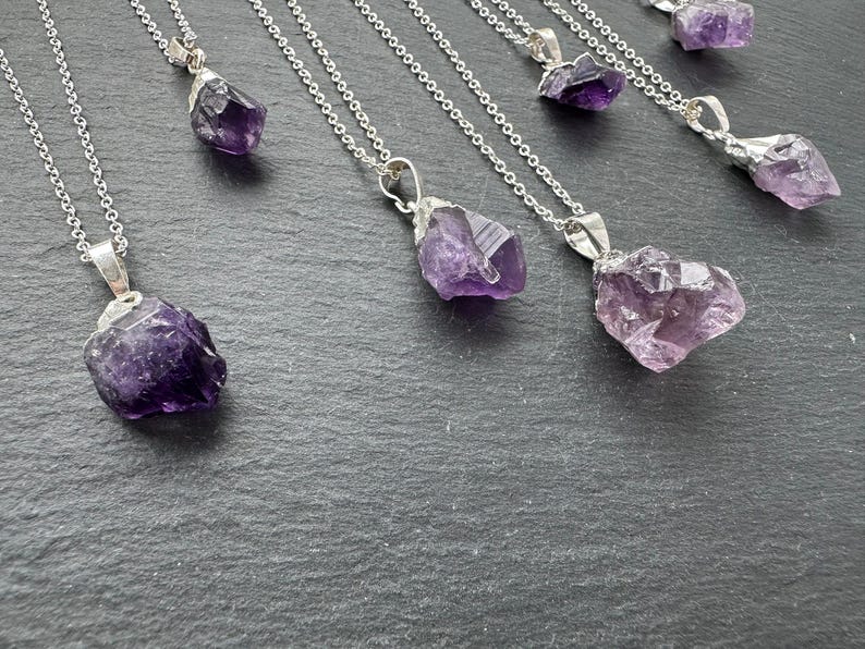 May include: A set of seven silver necklaces with amethyst crystal pendants. The crystals are raw and have a natural, irregular shape. The necklaces are all different lengths and the crystals are various sizes.