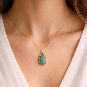 May include: A gold-colored necklace featuring a teardrop-shaped, faceted, green gemstone pendant. The pendant is framed in gold and hangs from a delicate gold chain. The necklace is displayed against a white background.