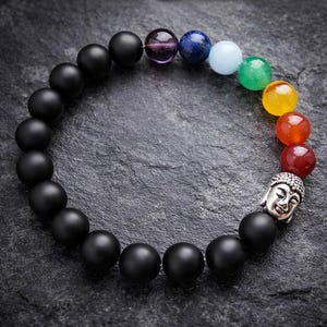 May include: A beaded bracelet featuring a silver Buddha head and a variety of colorful gemstones. The bracelet includes black onyx beads and a series of chakra stones in shades of purple, blue, green, yellow, orange, and red.