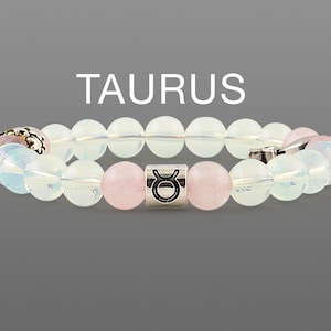 May include: A beaded bracelet featuring the word "TAURUS" in white text. The bracelet has a mix of clear, pink, and iridescent beads. A silver-colored cylindrical bead with the Taurus symbol is centered on the bracelet.