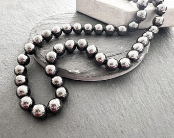 May include: A necklace composed of polished, dark gray, spherical beads. The beads are strung together, forming a continuous loop. The necklace is displayed on a gray, textured surface, with a portion resting on a dark gray, oval-shaped stone.