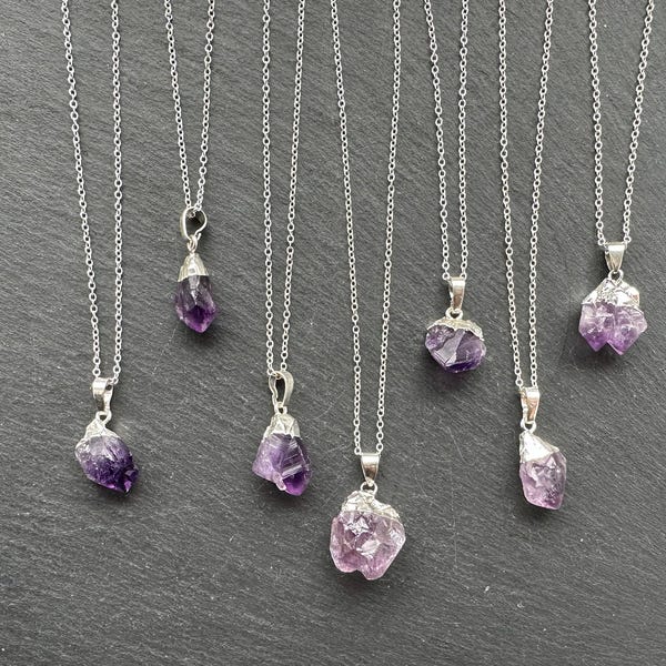 May include: Seven silver necklaces with amethyst crystal pendants. The amethyst crystals are all different shapes and sizes. The necklaces are all on a dark gray background.