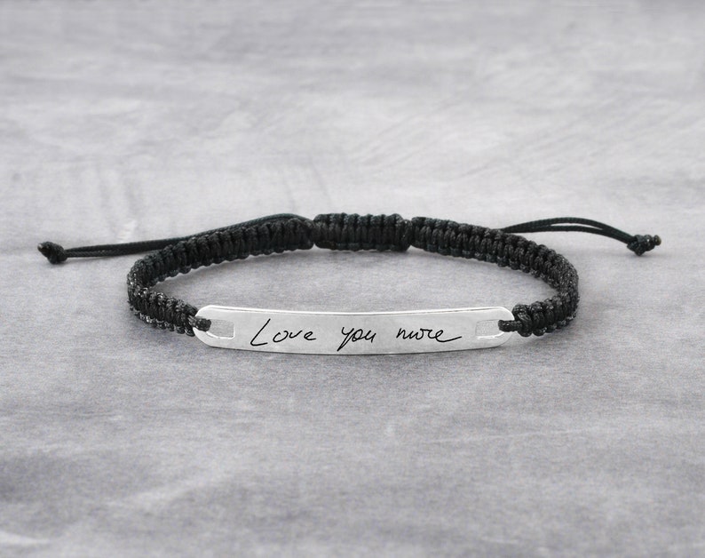 May include: A black braided cord bracelet with a silver bar charm engraved with the handwritten message "Love you more".