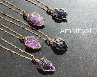 May include: Four gold necklaces with amethyst arrowhead pendants. The necklaces are layered on a dark gray surface. The word "Amethyst" is written in white text below the necklaces.