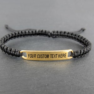 May include: A black braided bracelet with a gold metal tag that says "YOUR CUSTOM TEXT HERE".