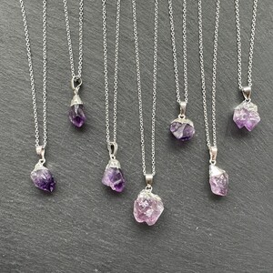 May include: Seven silver necklaces with amethyst crystal pendants. The crystals are all different shapes and sizes, and they have a purple and pink hue.